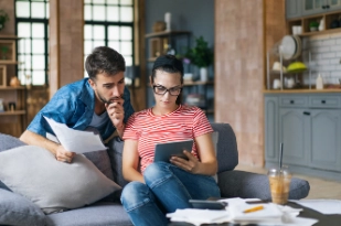 Couple calculant des factures à la maison à l’aide d’une tablette et d’une calculatrice. Jeune couple travaillant sur ordinateur tout en calculant ses finances assis sur le canapé. 