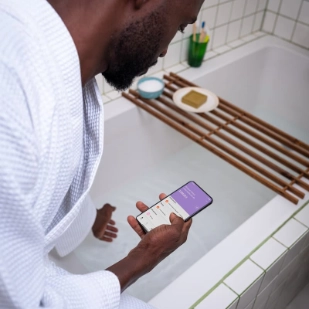 Homme en train de se faire couler un bain, teste la température avec le bout des doigts. Il tient un smartphone avec l'autre main.