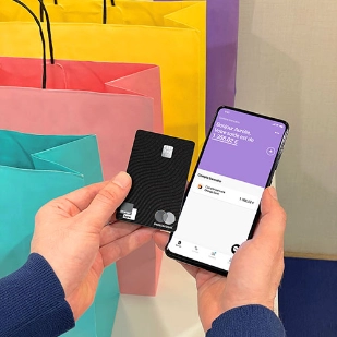 Mains qui tiennent une carte Premium et un mobile avec l'App Orange Bank ouverte, sur un arrière-plan de sacs de shopping.  