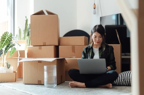 Jeune femme assise en tailleur au milieu de cartons, elle souscrit une assurance habitation locative depuis son ordinateur portable