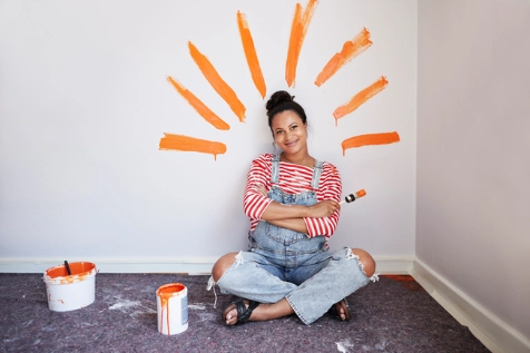 Une femme est assise sur le sol, de la peinture sur le mur forme des rayons autour d'elle, elle sourit et tient un pinceau