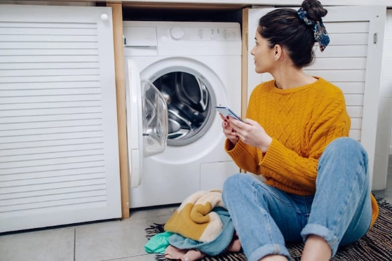 femme assise à côté d'une machine à laver le linge, elle tient un smartphone
