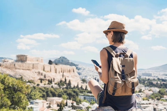 Une jeune femme consulte ses comptes depuis son smartphone à l'étranger, on aperçoit au loin le Parthénon