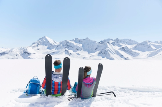 Un skieur et une skieuse sont assis dans la neige contre leurs skis, ils sont de dos et admirent le paysage
