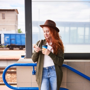 Femme rousse souriante  consulte son téléphone sur un quai en tenant son café